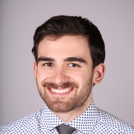 Smiling headshot, I have dark hair, a grey tie, and a white shirt with blue dots